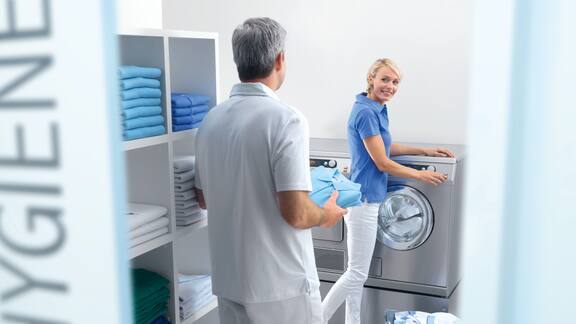 Työntekijä pesemässä lääkärinvastaanoton pyykkiä Miele Professional ‑pesukoneella. Hän on parhaillaan käynnistämässä konetta, kun lääkäri tulee noutamaan puhtaita työvaatteita.