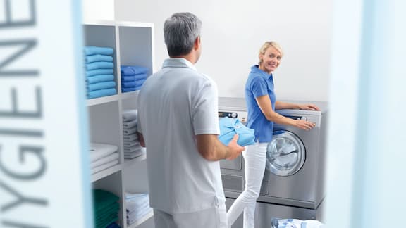 Medisinsk fagarbeider vasker klinikkens tekstiler i en Miele Professional-vaskemaskin. Hun slår på maskinen mens legen henter nyvaskede klær.