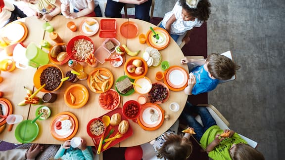 From bird’s-eye view: A breakfast table in a kindergarten with groceries on plastic plates, in plastic cups and with plastic cutlery. Children and nurses sitting around the table. 