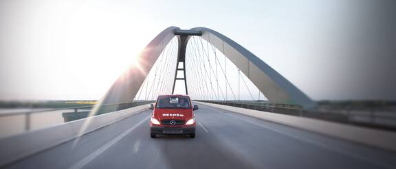 Piros színű autó halad a hídon háttérben a nappal.