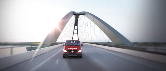 Rode auto rijdt over een brug met de zon op de achtergrond.