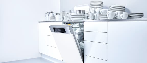 Range of Miele Professional Tank dishwasher: One throughput dishwasher and three tank dishwasher in a row, opened door equipped with baskets, glass and cutlery on grey background. In front another tank dishwasher with opened door and equipped.