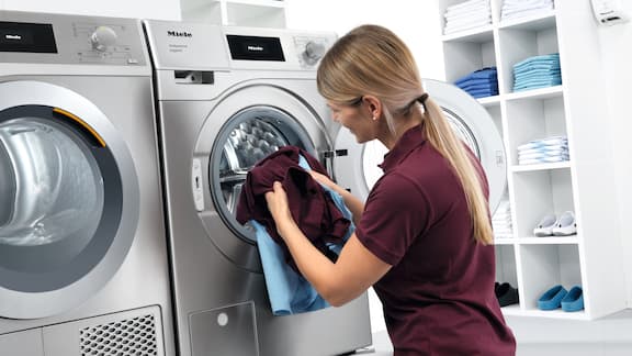 Mitarbeiterin kniet vor grauer Waschmaschine und befüllt diese mit Praxiskleidung.