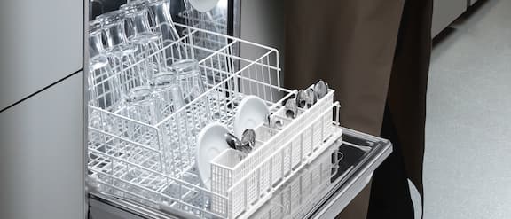 The basket of a tank dishwasher is pulled out onto the open dishwasher door. A waitress places crockery, cutlery and glasses in the basket of the dishwasher.