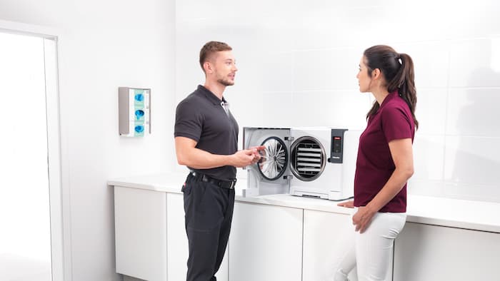 Male service technician in front of a steriliser talking to a female medical assistant at a practice.