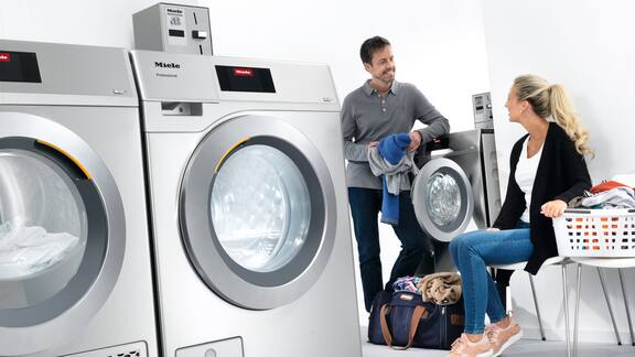 En kvinna och en man samtalar i ett tvättrum där grå maskiner tvättar och torkar tvätt.