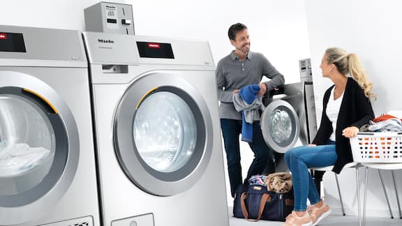 Vrouw en een man voeren een gesprek in een wasruimte, waar grijze machines wasgoed wassen en drogen.
