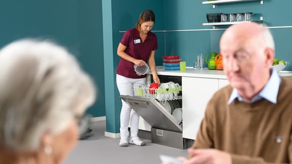 Pendant que les résidents seniors sont assis à table et jouent, une aide-soignante range le lave-vaisselle en arrière-plan.