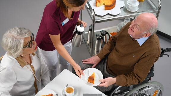 Eine Mitarbeiterin des Pflegeheims bringt zwei älteren Bewohnern Kuchen an den Tisch