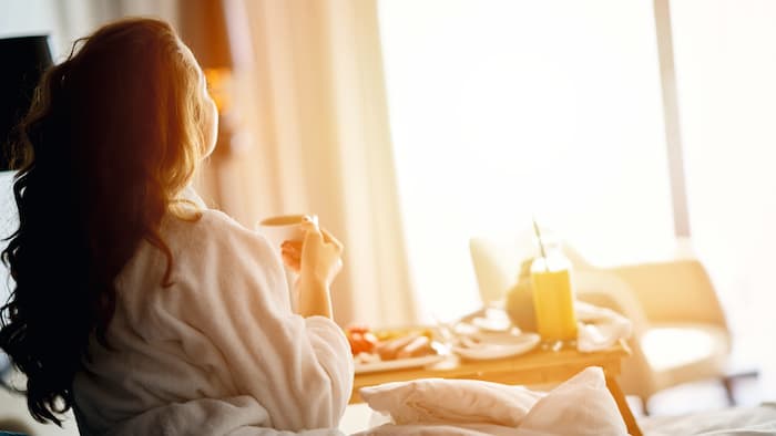 Hotelgästin beobachtet Sonnenaufgang aus Bett mit Kaffeetasse in Hand