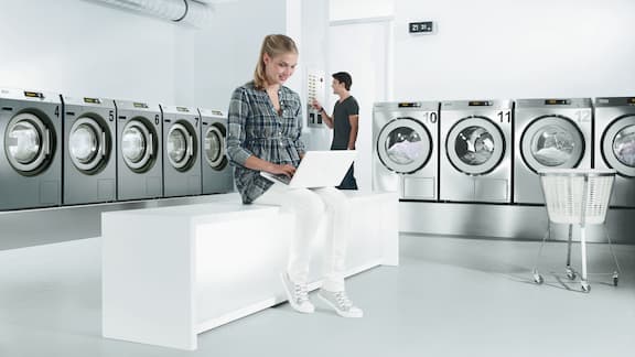 Une femme assise travaille sur un ordinateur portable dans une laverie automatique équipée de lave-linge et sèche-linge gris.