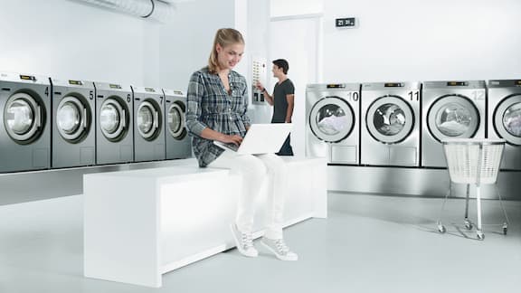 A woman sits with a laptop in a launderette with grey washing machines and tumble dryers.