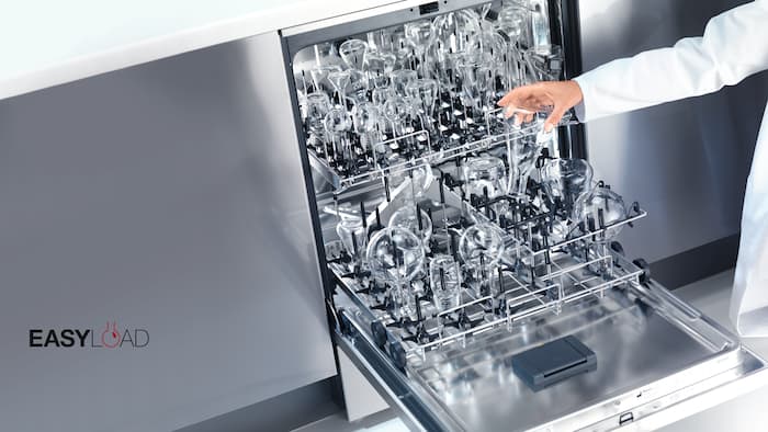 Nyitott laboratóriumi mosogatógép, amiben különféle laboratóriumi üvegek vannak a kosarakba helyezve.