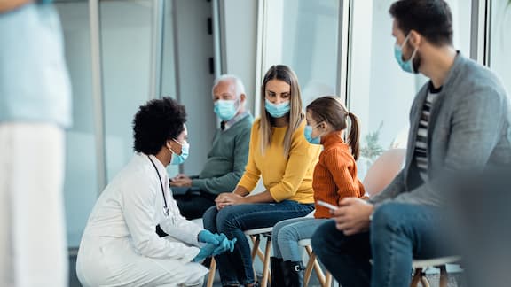 Um médico agacha-se em frente de pacientes na sala de espera. Três adultos e uma menina estão a aguardar pela consulta. Todos estão a usar máscaras.