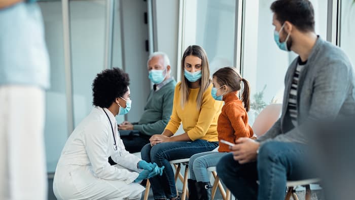 A doctor crouches in front of patients in the waiting room. Three adults and a little girl wait to go into the consulting room. All of them are wearing masks.