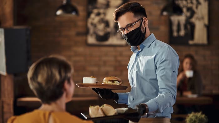 A waiter brings some burgers to a table. He is wearing a mask.