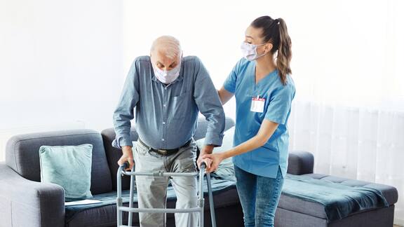 Egy női gondozó segít egy idős férfinak járókerettel járni. Mindkettő száj- és orrvédőt visel.