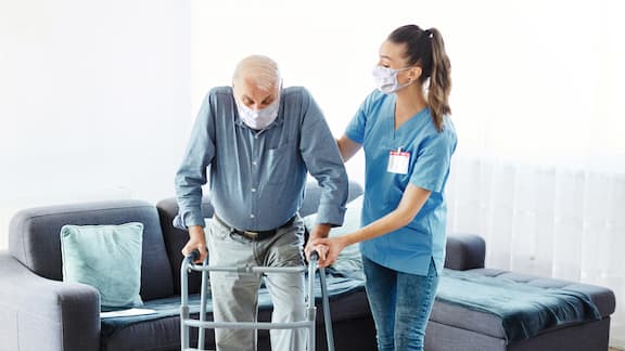 Een verpleegster ondersteunt een oudere man die met rollator loopt. Beiden dragen een mondmasker.