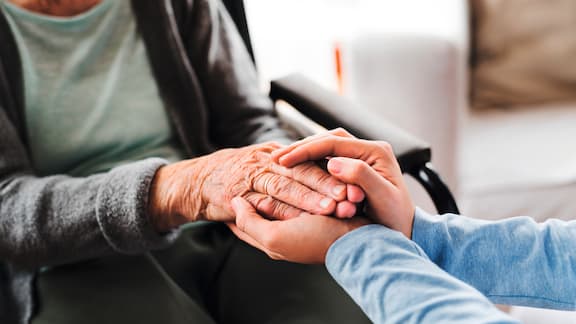 Two hands are holding the hand of an older person who is sitting in a chair.