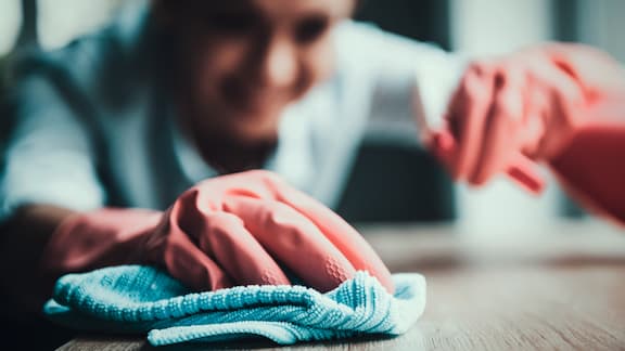 Een hand met roze handschoen maakt een oppervlak schoon met een blauwe reinigingsdoek.
