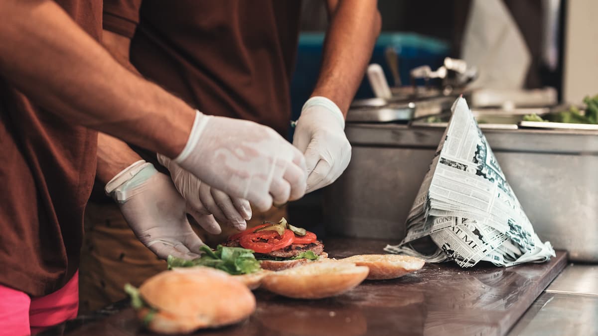 Hender med hvite hansker legger fyll på en burger på et kjøkken.