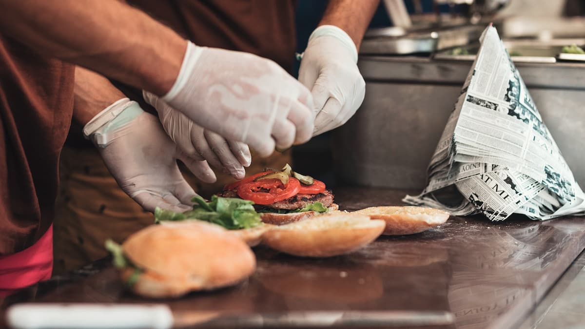 Hands in white gloves top burgers in the kitchen.