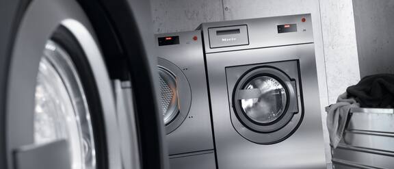 Reklámfotó sötét színű Miele Professional mosógépről ezüst ruháskosárral.