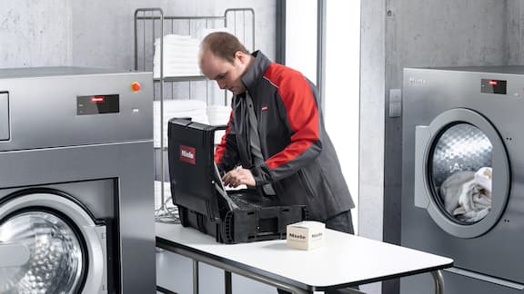 Een technicus staat voor zijn gereedschapskoffer en een doos onderdelen, vlakbij een wasmachine en droger