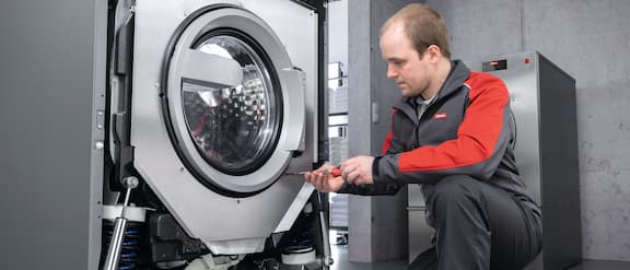 A service engineer is repairing a washing machine with the front removed.