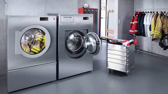 Fire station laundry room with various fire brigade textiles hanging on a rail as well as a washing machine and tumble dryer.