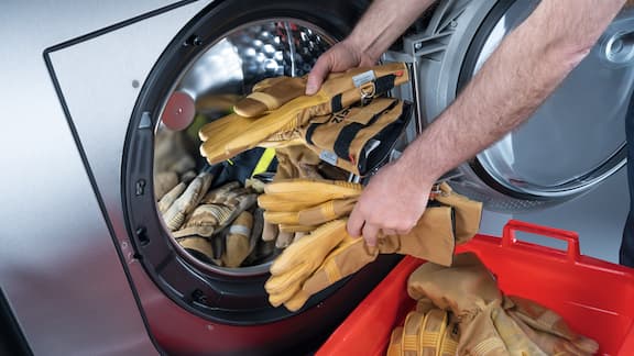 Mãos masculinas carregam uma máquina de lavar roupa Miele Professional com luvas de bombeiro.