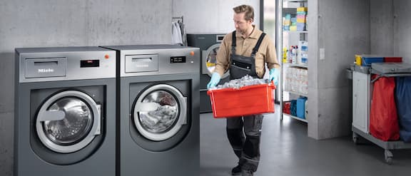 Bild einer Wäscherei mit zwei unterschiedlich großen Miele-Waschmaschinen und einer Person mit schmutziger Wäsche.
