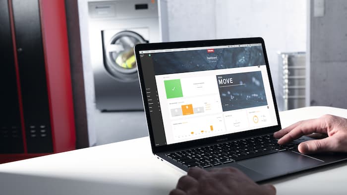 A laptop displaying the Miele MOVE platform is positioned on a white desk. A Miele Benchmark Performance Plus washing machine can be seen in the background.