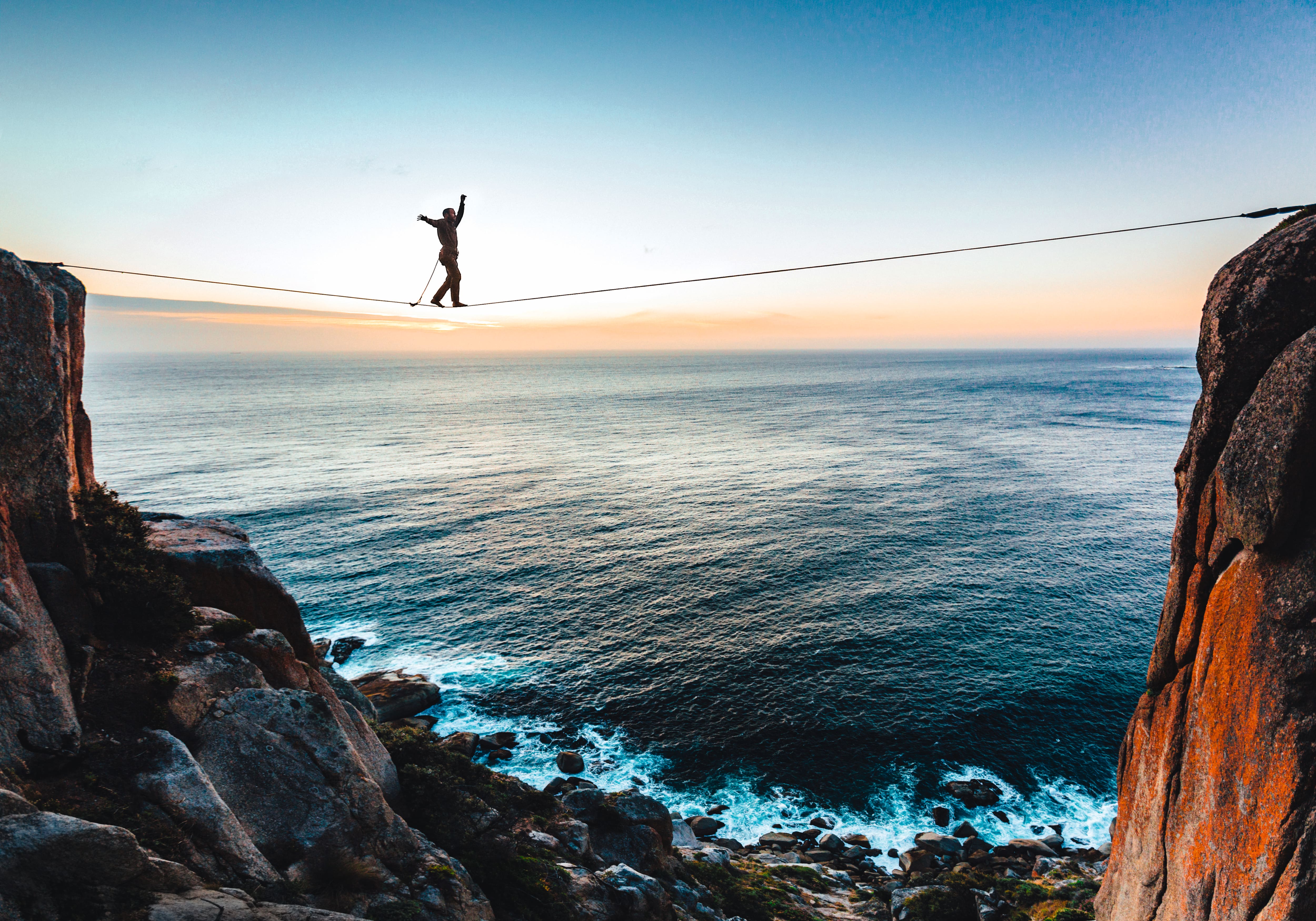 Slackliner balanciert auf einem Seil zwischen zwei Gipfeln und dem Meer im Hintergrund.]