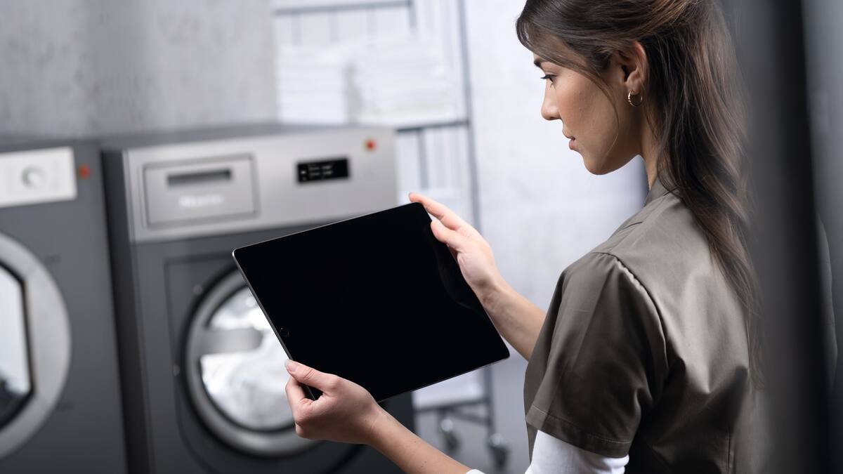 Egy nő egy tabletet tart a kezében, és egy szürke mosógép előtt áll.
