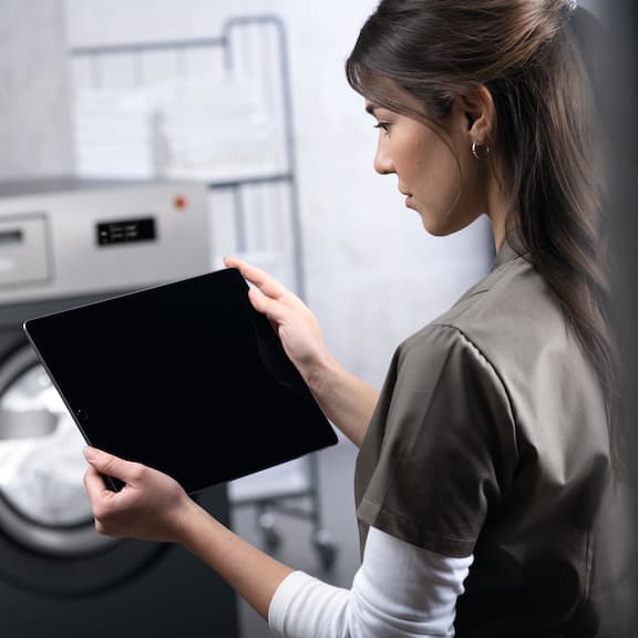 Une femme, tablette en main, se tient devant un lave-linge gris.