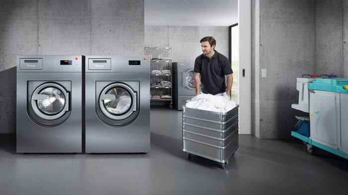 Two Benchmark Performance Plus washing machines are standing next to each other in the laundry room. In the background, a care worker dressed in white is standing next to a Benchmark Performance Plus tumble dryer and is sorting laundry on a rack.