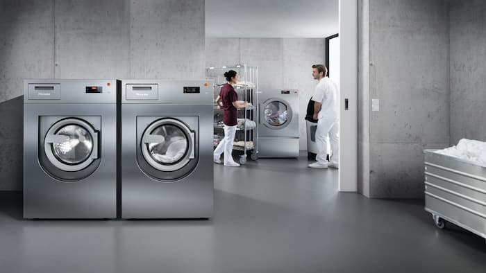 A restaurant worker carrying a laundry basket full of tea towels is entering the laundry room where there is a tumble dryer and a washing machine from the Benchmark Performance Plus series.