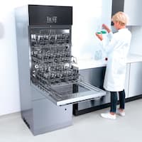 Lavadora de laboratorio SlimLine abierta y cargada situada en una sala de limpieza donde una empleada de laboratorio sostiene dos matraces Erlenmeyer en la mano.