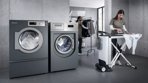 A woman irons a shirt with the Miele steam ironing system. There is a grey washing machine and tumble dryer in the background.