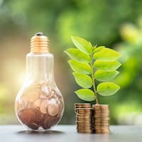 Light bulb filled with money coins next to a pile of money coins from which a plant grows in front of a green background