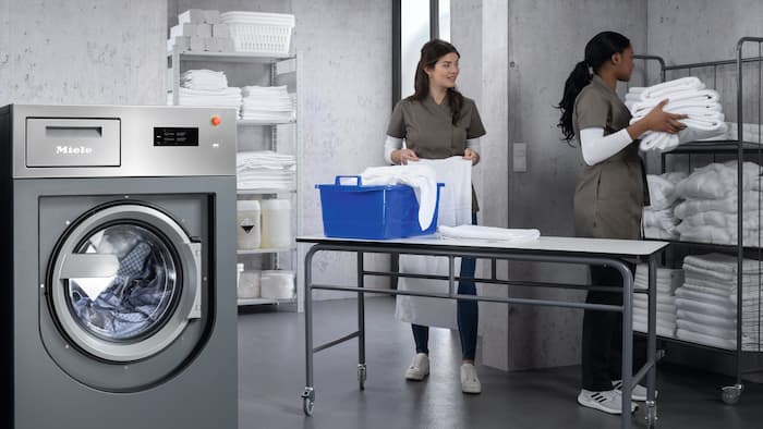 Modern laundry facility with Miele Professional Benchmark washing machine in the foreground. 2 employees in grey workwear are folding and sorting fresh towels.