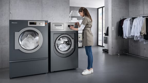 A woman operates a grey tumble dryer in a washroom. Next to it is a grey washing machine.