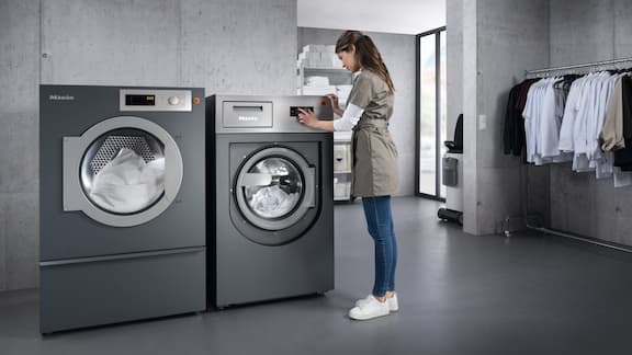 Une femme utilise un sèche-linge gris dans une buanderie. À côté, un lave-linge gris.