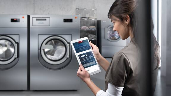 Eine Wäschereiangestellte steht in der Inhouse Wäscherei eines Pflegeheims und schaut auf ein Tablet auf dem Miele MOVE zu sehen ist. Im Hintergrund stehen die Miele Professional Benchmark Trockner und Waschmaschinen. 