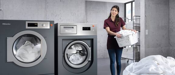 Een schoonmaakhulp in een hotel draagt een wasmand met handdoeken de wasruimte in. Naast de hulp staan de Benchmark-machines.