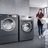 A hotel housekeeper is carrying a laundry basket of towels into the laundry room. The Benchmark machines are next to her.