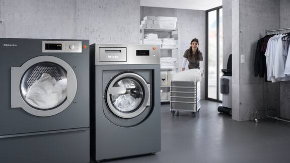 Wäschereimitarbeiterin schiebt Wäschewagen zu Miele Professional Gewerbewaschmaschine und Gewerbetrockner 