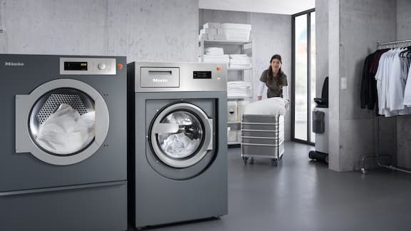 Laundry worker moves laundry trolley to Miele Professional commercial washing machine and commercial tumble dryer