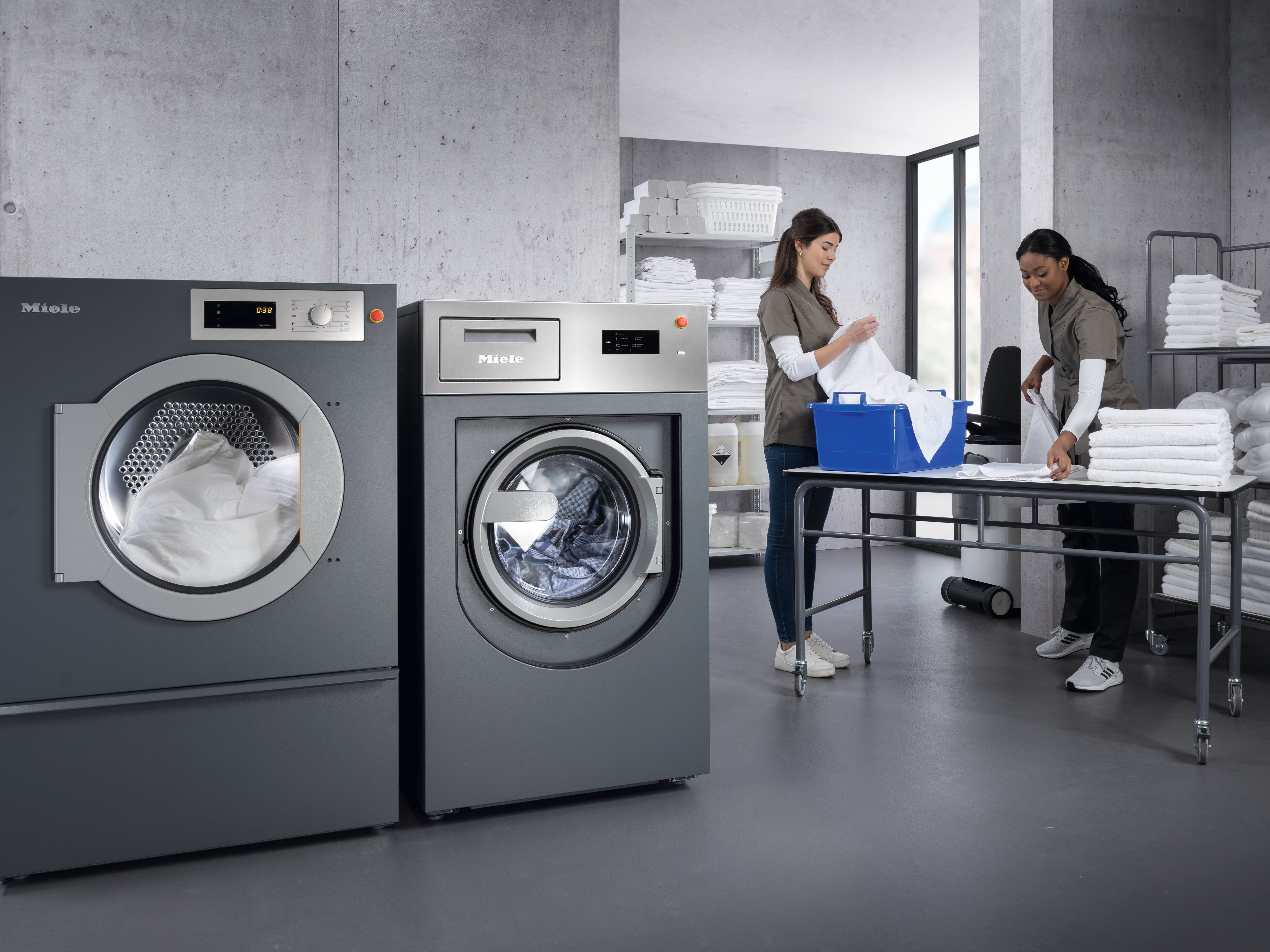 Dwie osoby pracują w jasnej pralni z urządzeniami przemysłowymi, składając ręczniki i dozując środki piorące.
